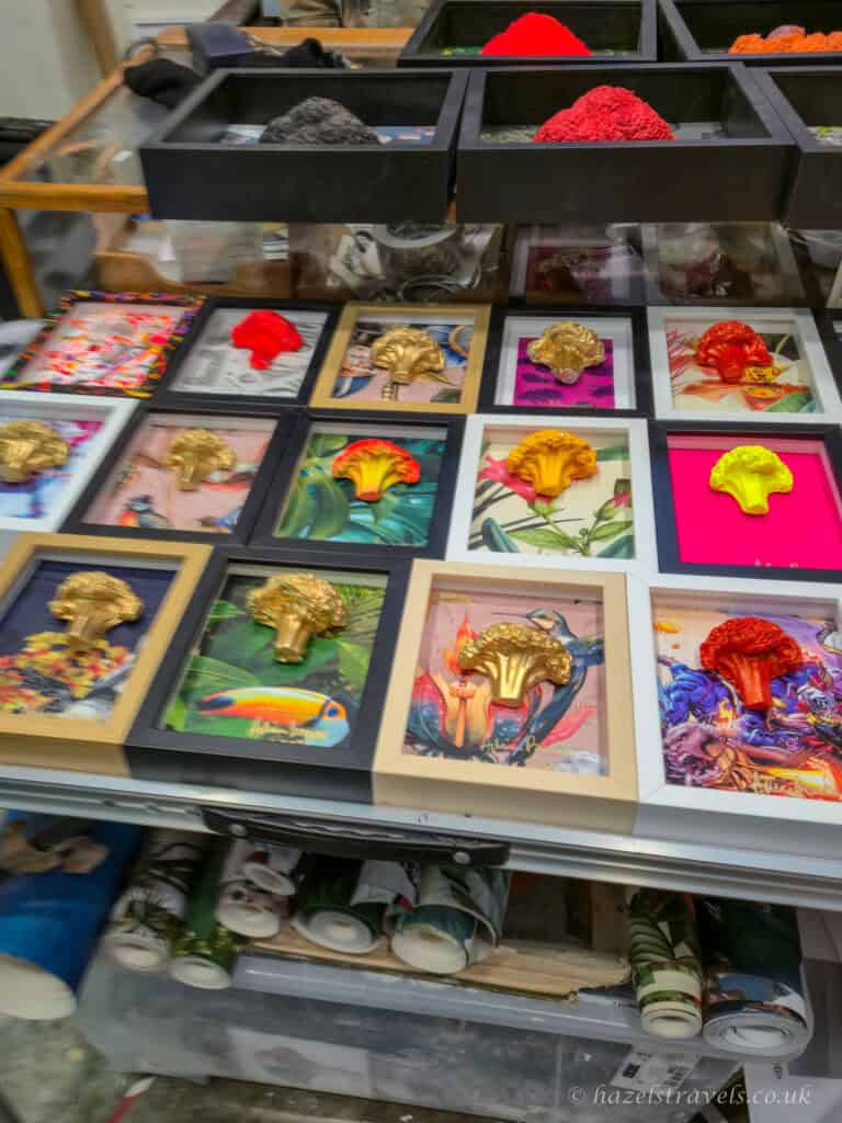 Colourful framed broccoli art displayed on a market stall, featuring gold and brightly painted broccoli sculptures mounted on vibrant illustrated backgrounds