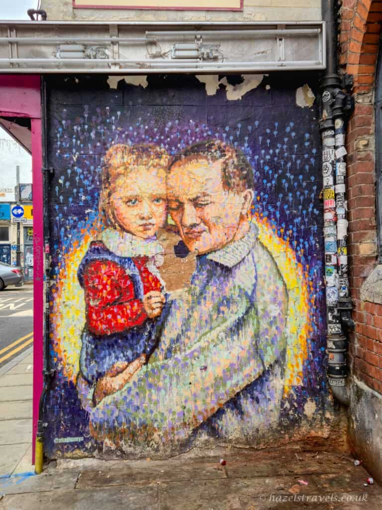 Street art mural in Shoreditch, London, depicting a man holding a young child, painted in layered dots of blue, yellow and warm tones on a weathered brick wall