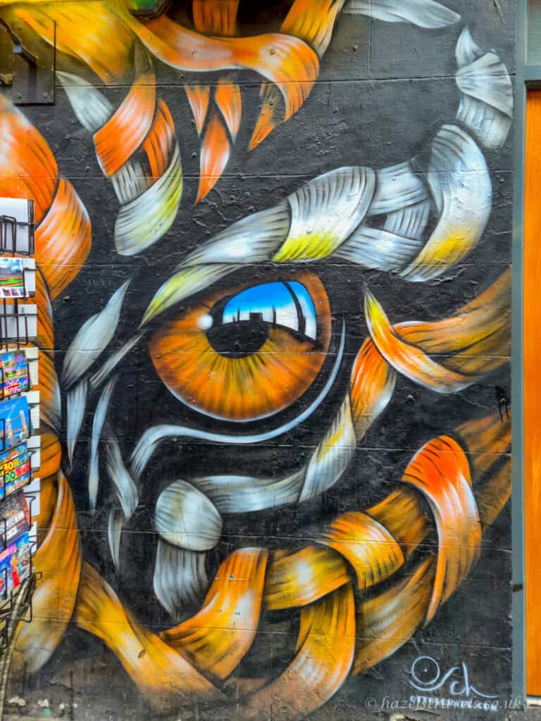 Street art mural in Shoreditch, London, featuring a striking close-up eye surrounded by swirling feather-like shapes in orange, gold and white painted on a dark wall