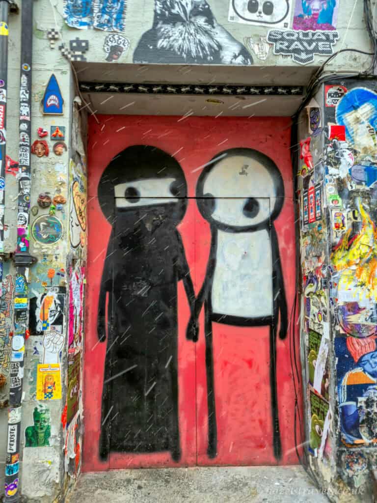 Street art doorway in Shoreditch, London, featuring two simple cartoon-style figures painted in black and white on red doors, surrounded by a dense collage of stickers and graffiti