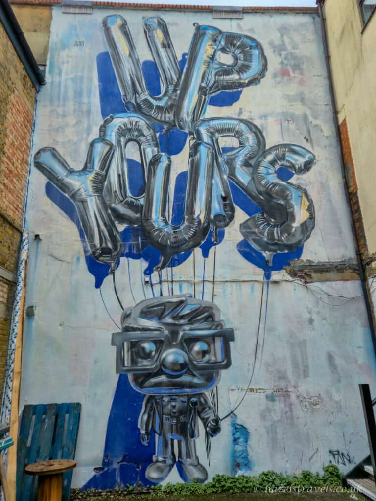 Street art mural in Shoreditch, London, reading “UP YOURS” in metallic balloon-style lettering above a cartoon figure holding the strings, painted on a tall urban wall