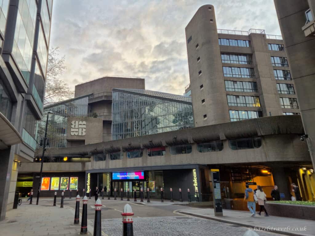 Exterior view of the Barbican Centre in London, showing grey Brutalist concrete towers and a pale glass conservatory structure, with dark entrance signage and a cloudy blue-grey sky above.