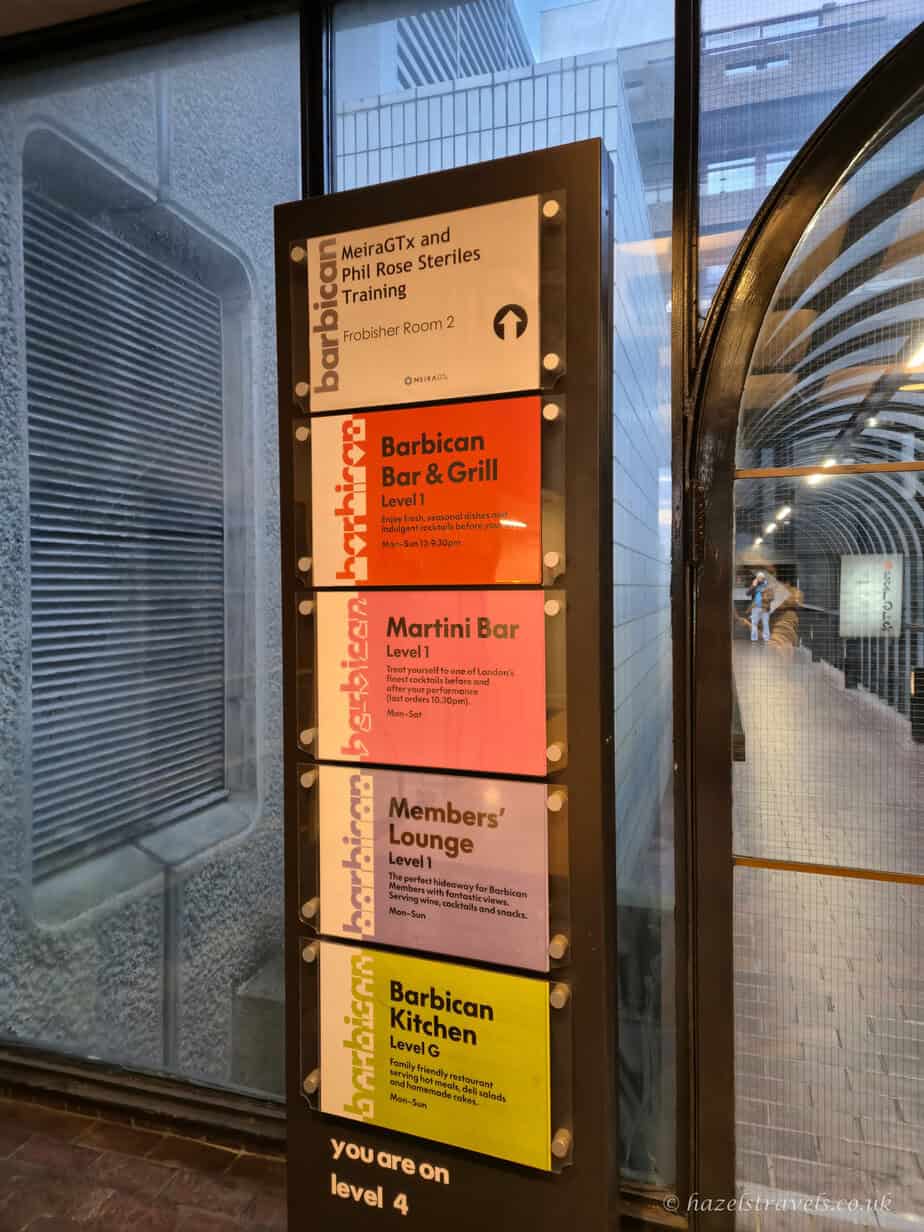 Colour-coded directory sign inside the Barbican Centre, with stacked panels in orange, red, yellow, and pink listing bars and venues, set against grey concrete walls and a curved glass corridor.