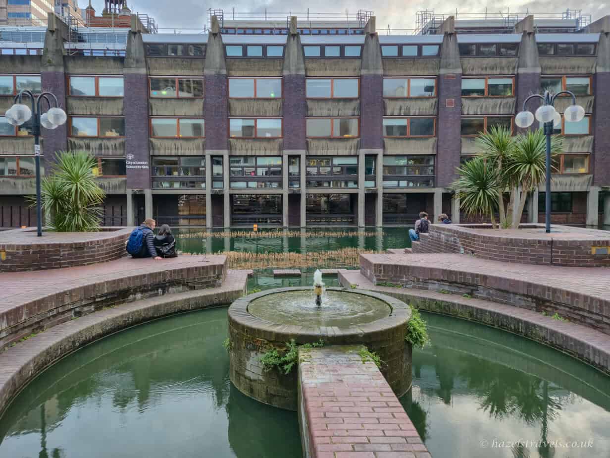 Circular green water feature with a small central fountain, red-brown brick walkways and steps, people sitting along the edges, and grey and brown Brutalist buildings under a cloudy pale grey sky.