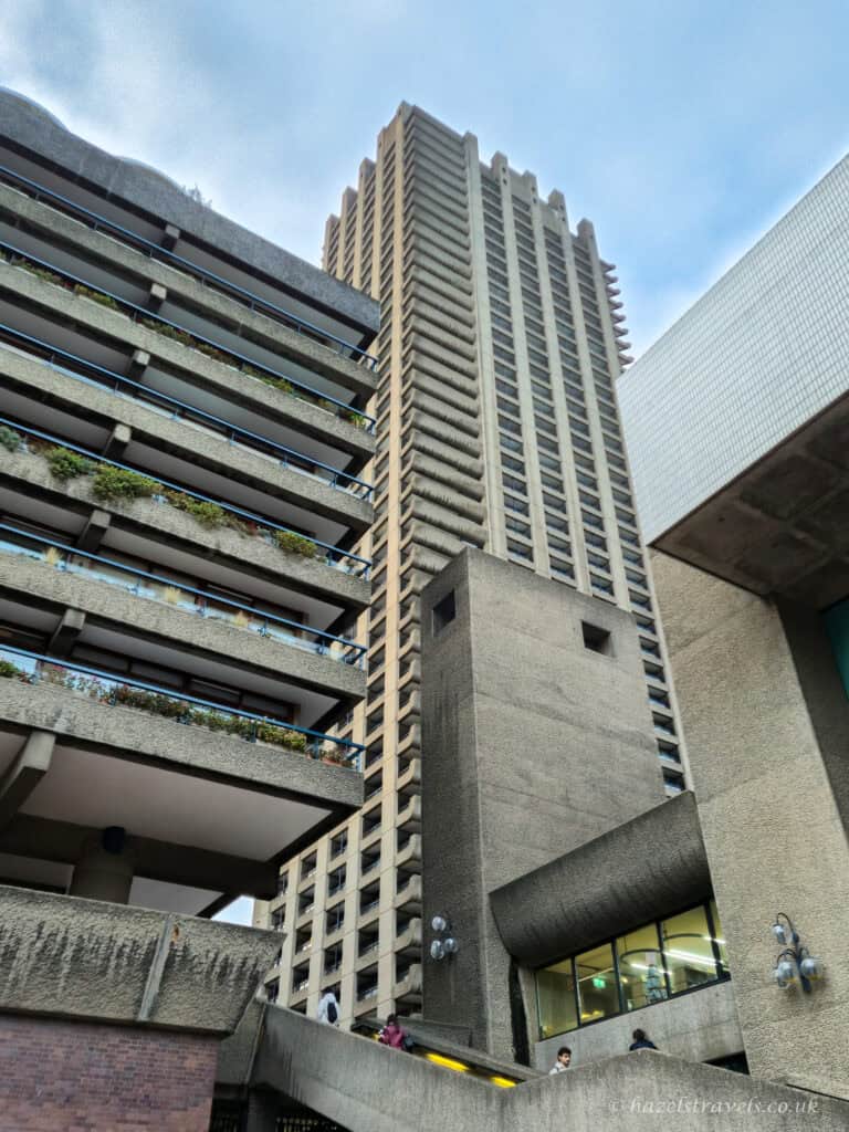 Tall grey Brutalist tower viewed from below, with layered concrete balconies, small windows, and surrounding angular buildings against a pale blue sky.