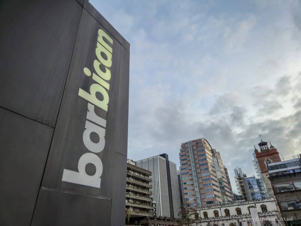 Barbican Centre sign on a dark exterior wall, with Brutalist towers and surrounding London buildings under a cloudy sky.