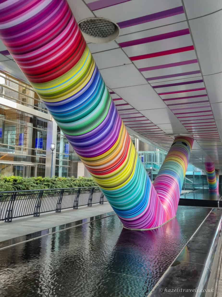 Vibrant rainbow-striped sculpture wrapping around large pillars under a covered walkway in Canary Wharf, reflecting in the shallow water feature below with modern buildings in the background.