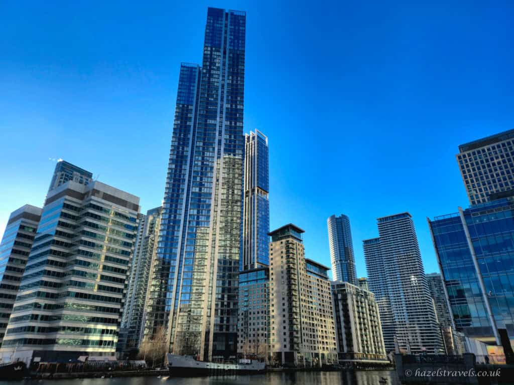 Tall glass skyscrapers and waterfront apartment buildings in Canary Wharf, reflecting the sunlight under a bright blue sky. The scene is viewed from across the water, with calm reflections and modern architecture dominating the skyline.
