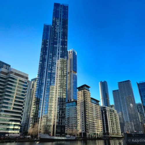Tall glass skyscrapers and waterfront apartment buildings in Canary Wharf, reflecting the sunlight under a bright blue sky. The scene is viewed from across the water, with calm reflections and modern architecture dominating the skyline.