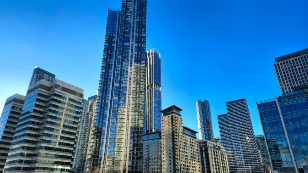 Tall glass skyscrapers and waterfront apartment buildings in Canary Wharf, reflecting the sunlight under a bright blue sky. The scene is viewed from across the water, with calm reflections and modern architecture dominating the skyline.