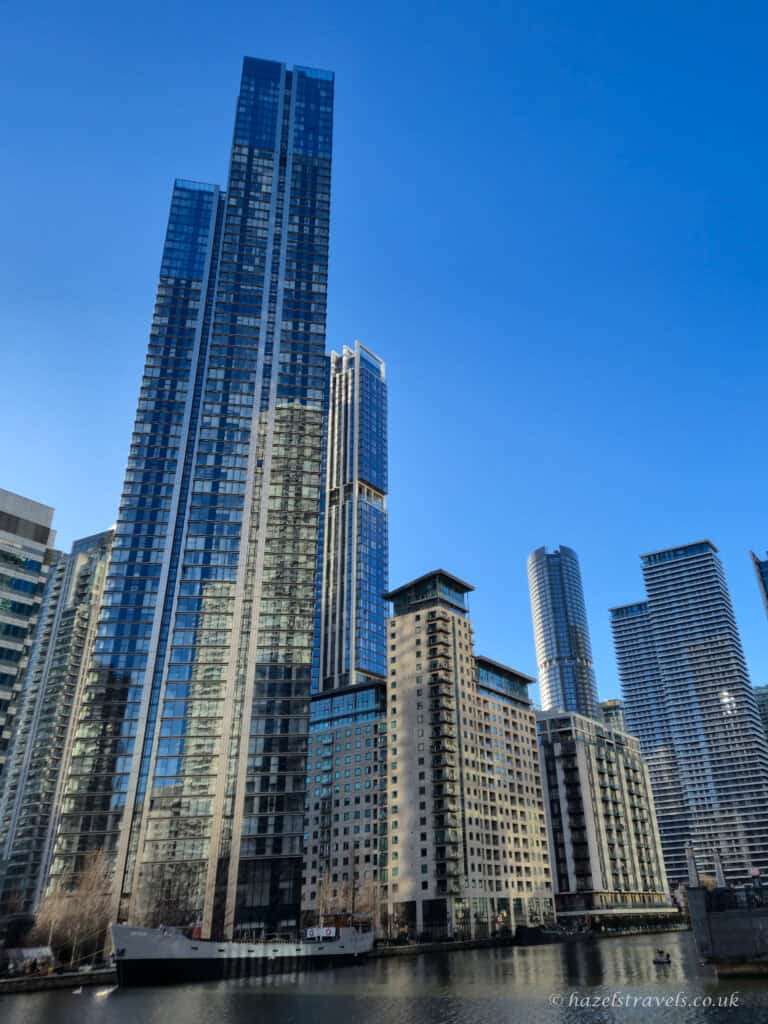Tall modern skyscrapers in Canary Wharf reflected in still dock water, with a clear blue sky above. The central tower is glass-fronted and surrounded by a mix of high-rise offices and residential buildings.