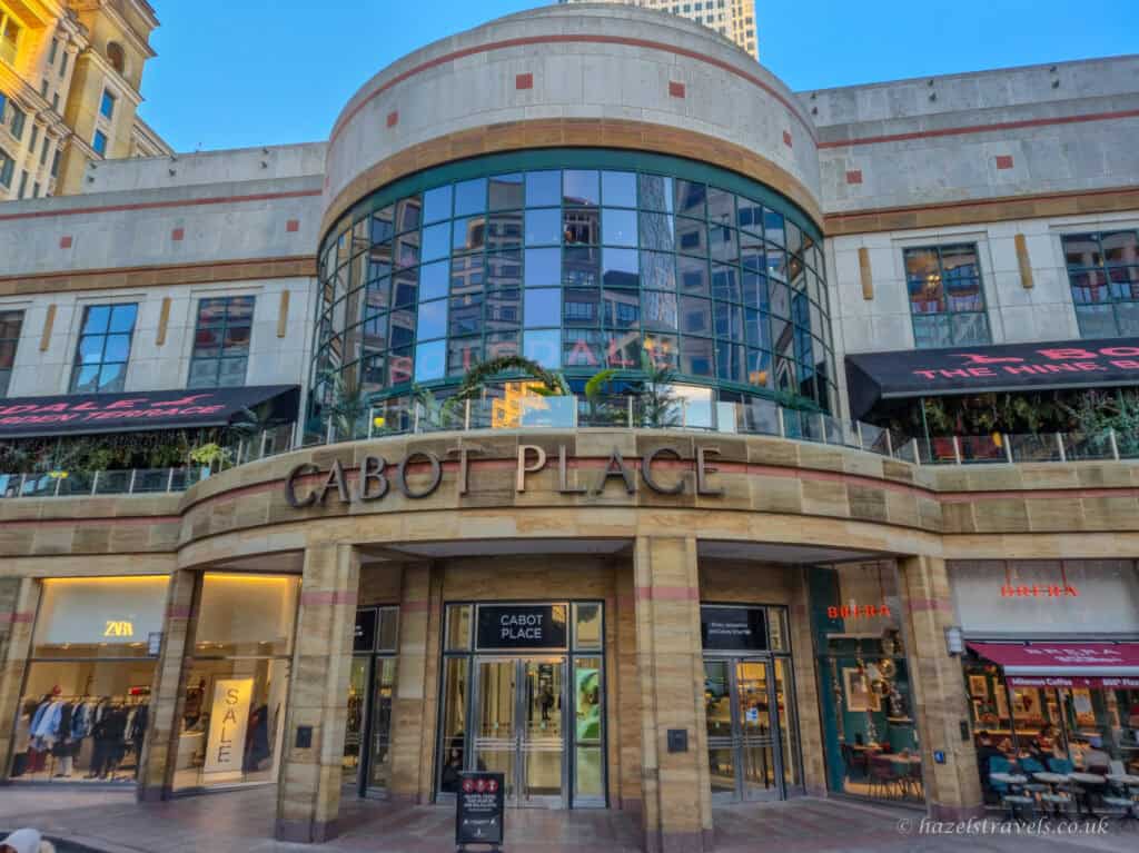 Entrance to Cabot Place shopping centre in Canary Wharf on a clear day, showing a curved stone and glass facade with large arched windows, shopfronts including Zara and Brera, and reflections of surrounding skyscrapers in the upper glass panels.