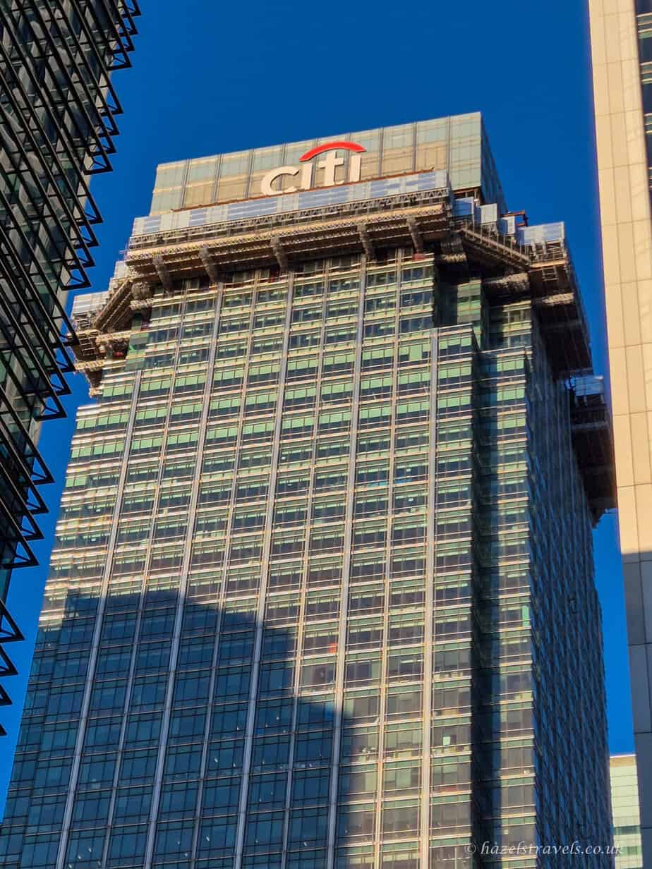 Tall glass office tower in Canary Wharf with the Citibank logo at the top, captured on a clear day with deep blue sky and strong shadows from nearby buildings.