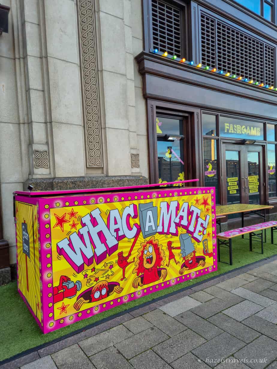Bright pink and yellow 'Whac-A-Mole' game outside Fairgame in Canary Wharf, with colourful cartoon graphics and lights above the entrance to the retro-style bar and arcade venue.