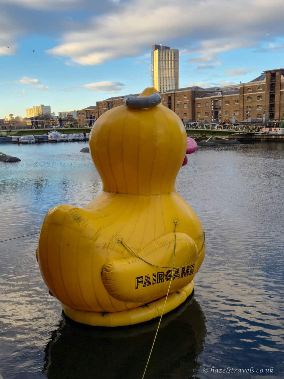 Giant inflatable yellow rubber duck with a Fairgame logo floating on the water in Canary Wharf, facing brick buildings along the dock under a partly cloudy evening sky.