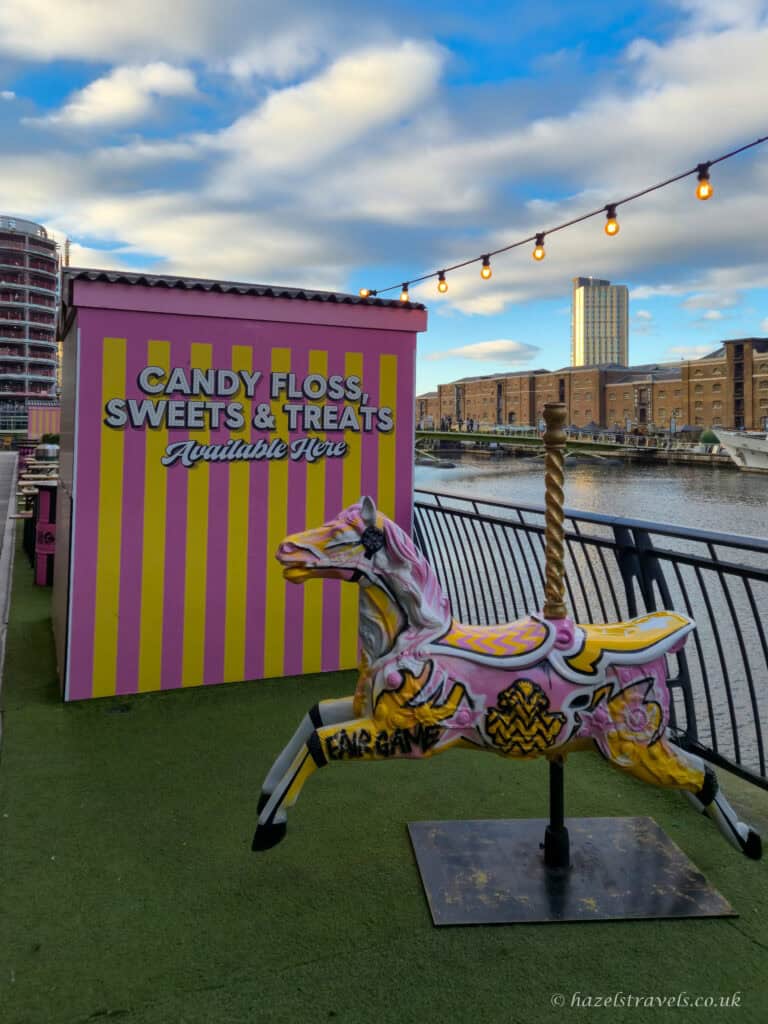 Candy floss and sweets stall at Fairgame in Canary Wharf, painted in pink and yellow stripes with a colourful carousel horse sculpture in front. The setup overlooks the dock, with festoon lights overhead and modern buildings in the background under a partly cloudy sky.