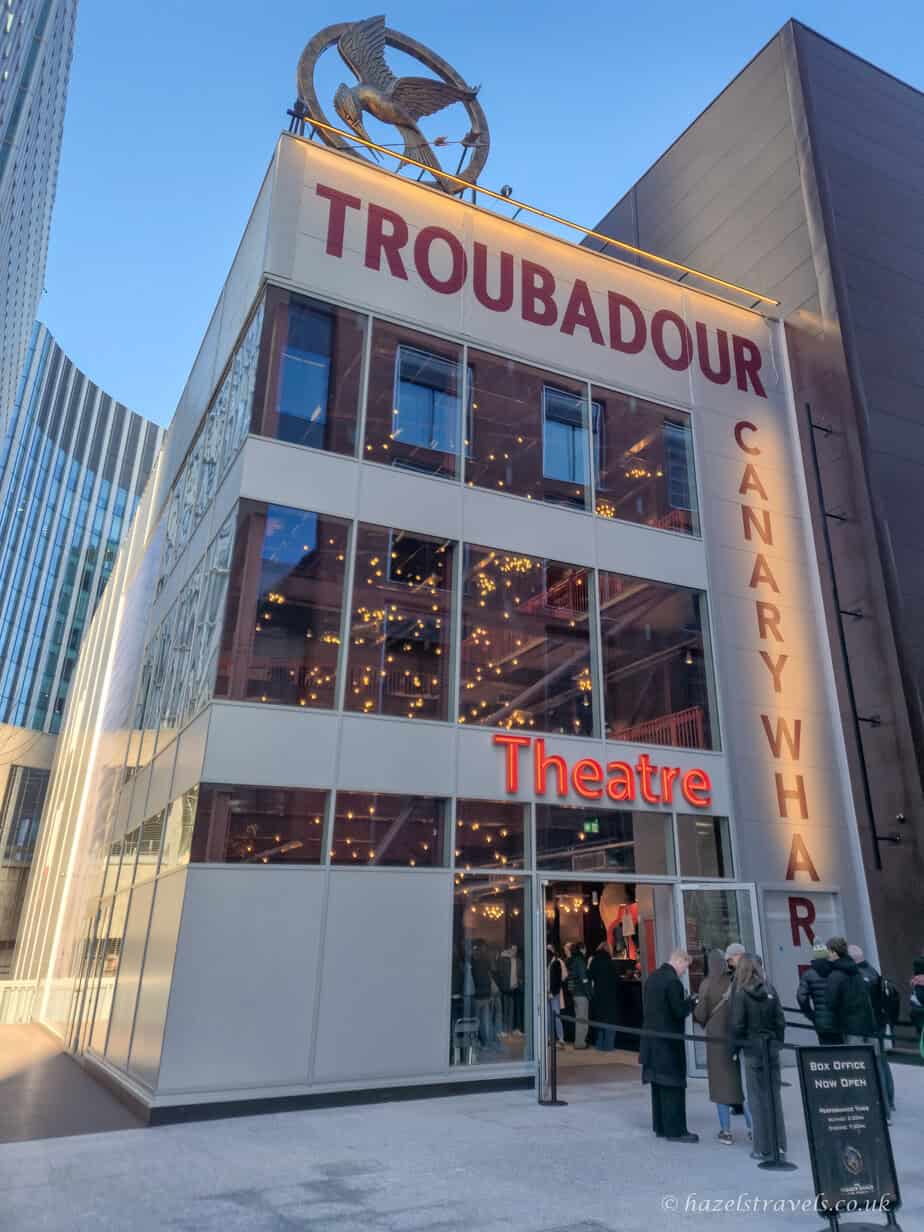Exterior of the Troubadour Theatre in Canary Wharf, a modern white and red building with large windows and a glowing red 'Theatre' sign. People are gathered at the entrance under a clear blue sky, and string lights are visible through the windows.