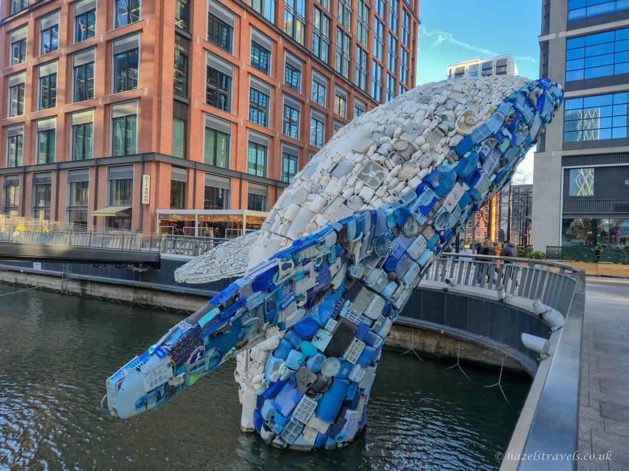 Large whale sculpture made from recycled plastic waste, including white containers and blue household items, emerging from the water in a dockside area of Canary Wharf. The backdrop features modern red-brick and glass buildings, with pedestrians and a restaurant visible nearby under a clear blue sky.