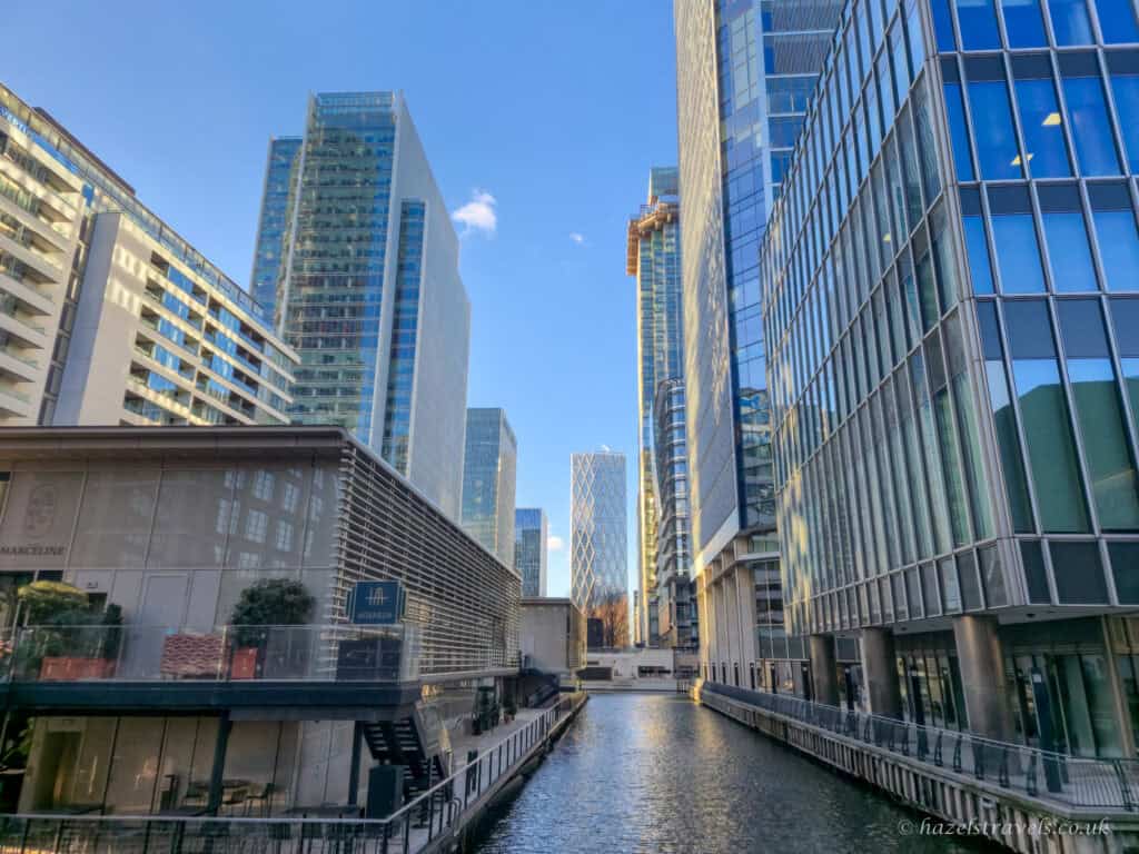 A calm view along a narrow canal in Canary Wharf, flanked by modern glass office towers reflecting blue sky, with water leading the eye towards distant high-rises.