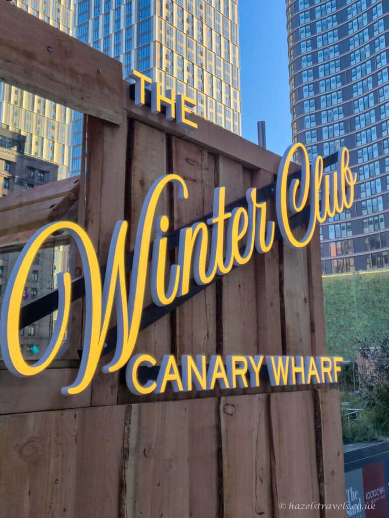 Wooden sign with glowing yellow script text reading 'The Winter Club Canary Wharf', set against modern high-rise buildings on a clear day. The festive installation hints at seasonal activities in the heart of the financial district.