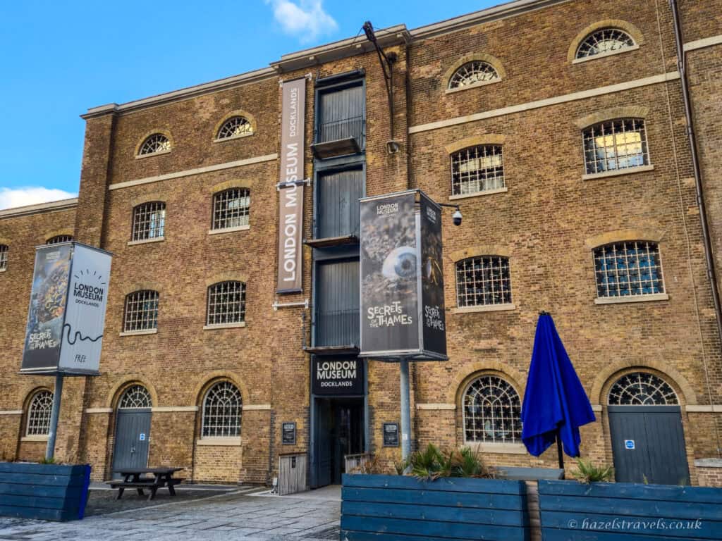 The London Museum of Docklands on a bright day, with a tall brown-brick warehouse building, arched windows with white bars, museum banners hanging on the facade, and a blue sky with scattered clouds above.