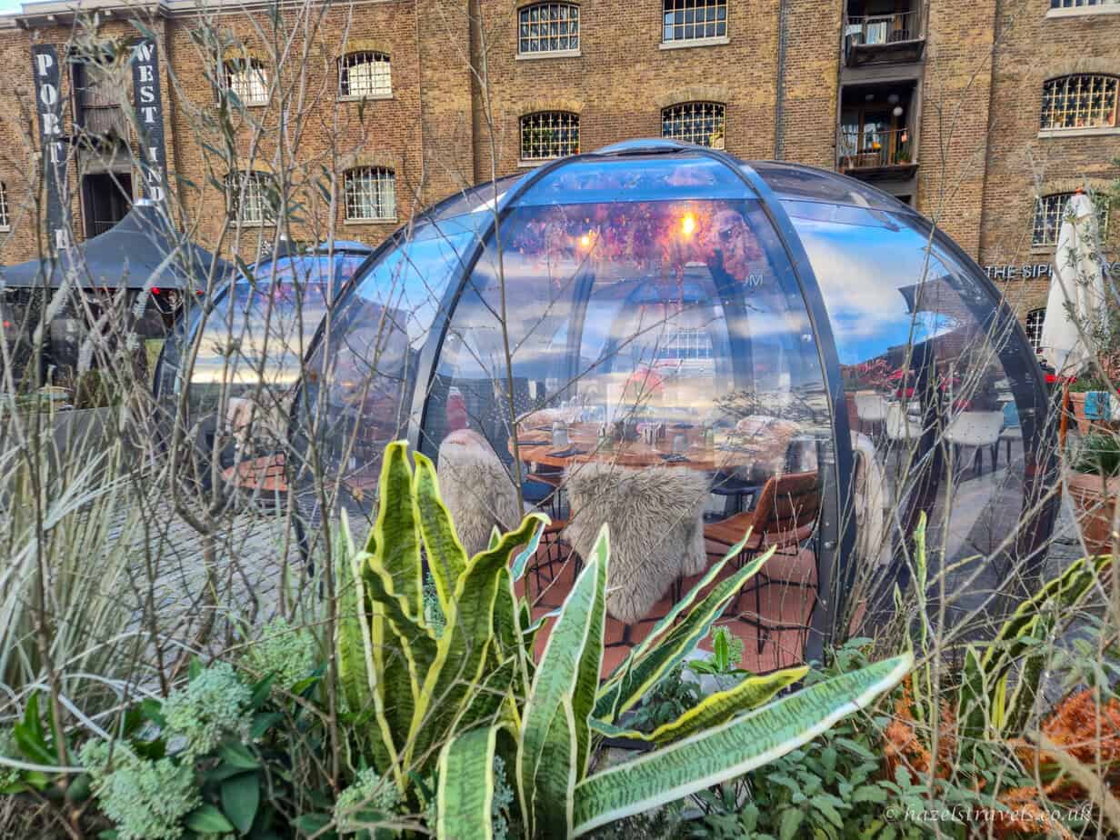 Cosy transparent dining igloo with soft lighting and fur-lined chairs at The Sipping Room in Canary Wharf, surrounded by greenery and set against a backdrop of historic brick buildings.
