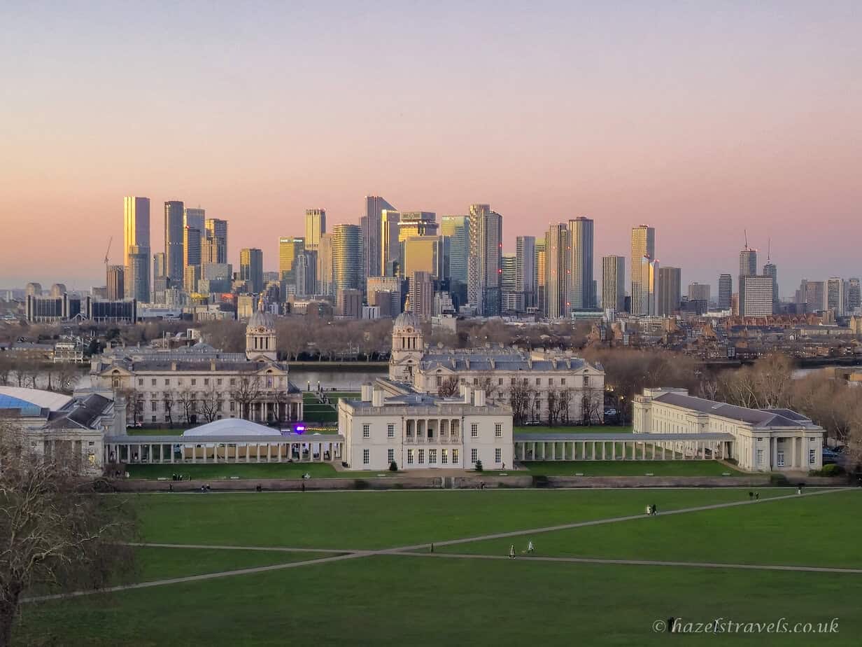 Sunset view from Greenwich Park with wide green lawns in the foreground, the cream and white Old Royal Naval College below, and the Canary Wharf skyline glowing gold and silver under a soft pink and pale blue evening sky in London