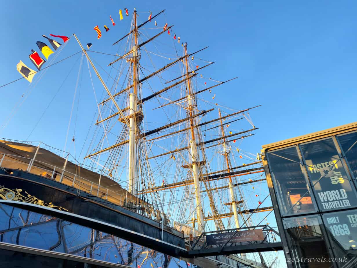 Towering masts and tangled rigging of the Cutty Sark decorated with colourful signal flags, rising against a clear bright blue sky above the dark hull and glass structure at Maritime Greenwich, London
