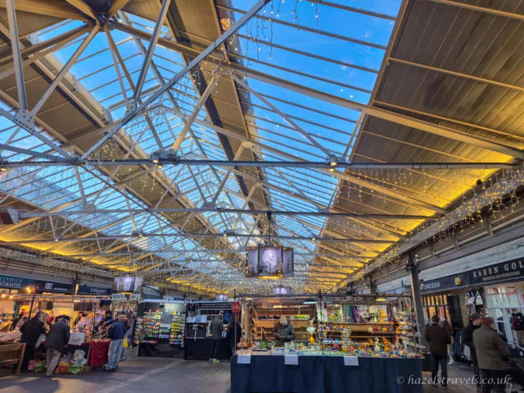 Interior of Greenwich Market with a glass and steel roof glowing with warm yellow fairy lights, deep blue evening sky visible above, and colourful craft stalls and shoppers below in soft golden winter light, London