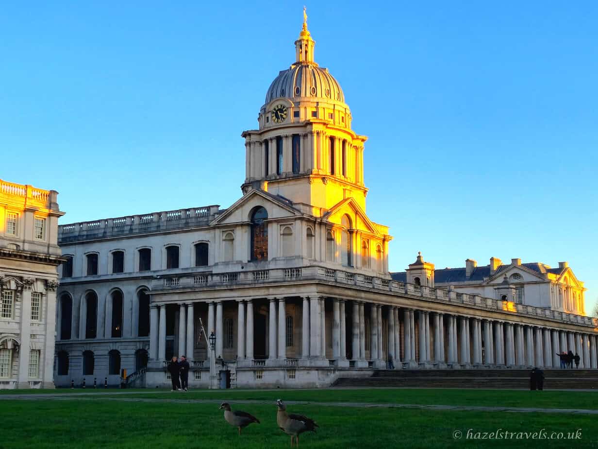 Long cream and pale yellow façade of the Old Royal Naval College in Greenwich bathed in warm golden sunset light, with deep blue winter sky, bare tree branches overhead, and dark green grass in the foreground, London