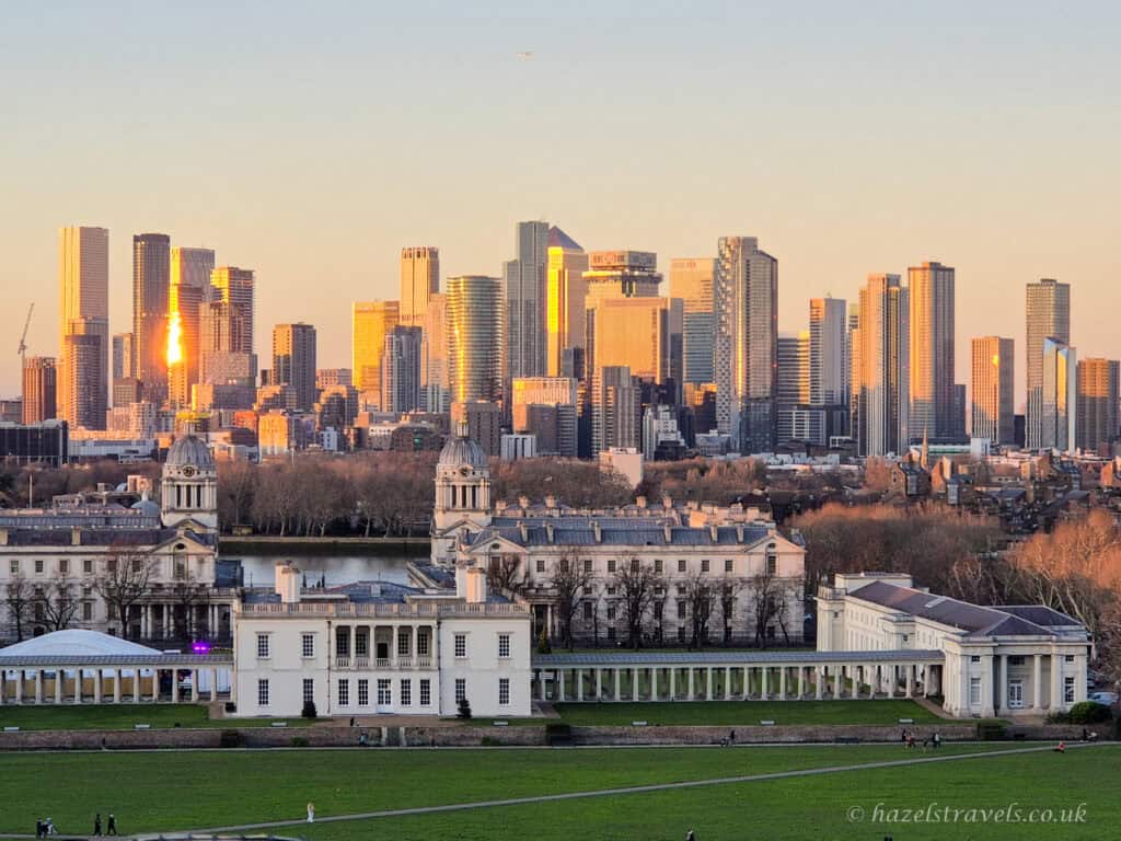 View from Greenwich Park at sunset with pale green grass in the foreground, cream and white neoclassical buildings of the Old Royal Naval College below, and the Canary Wharf skyline glowing gold and amber in warm evening light against a soft pastel blue sky in London