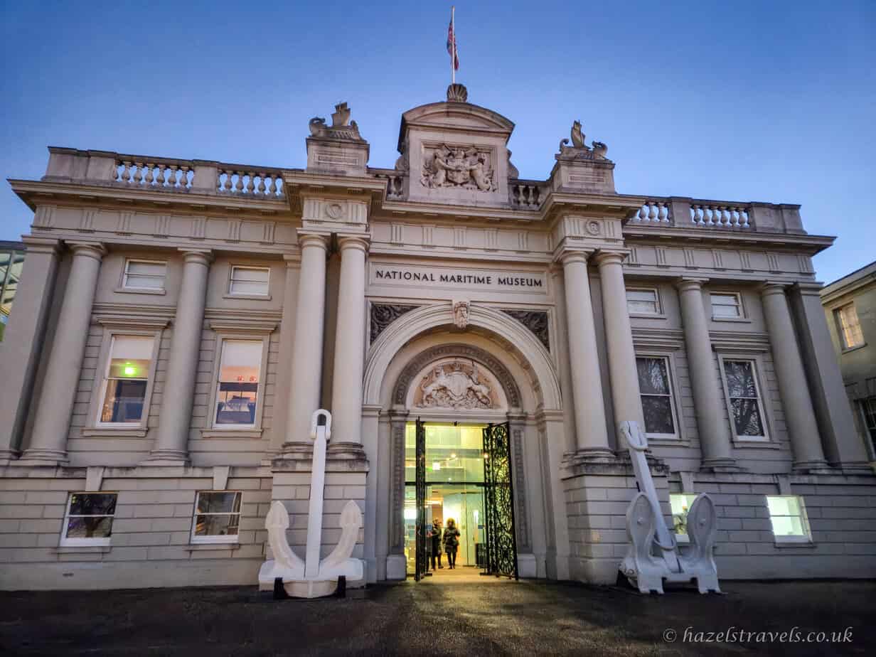 Front façade of the National Maritime Museum in Greenwich at dusk, with pale stone columns, carved archway, white anchor sculptures flanking the entrance, warm interior lights glowing through open doors, and a clear deep blue evening sky, London