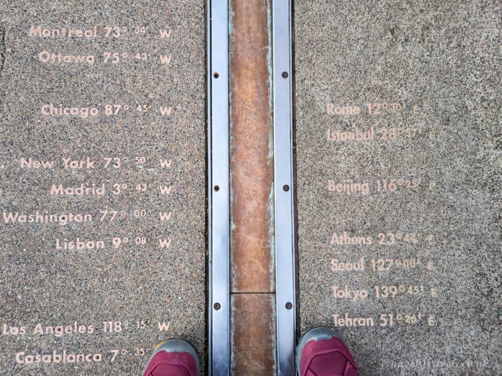 The Prime Meridian line at the Royal Observatory in Greenwich marked by a bronze strip set into grey stone paving, with city names and longitudes etched on either side and a pair of burgundy shoes standing astride the line, London