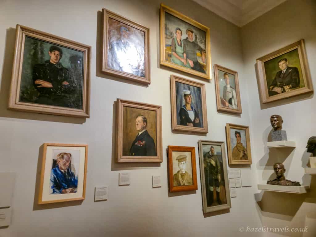 Gallery wall inside the Queen’s House in Greenwich displaying framed portrait paintings in muted greens, browns and creams, depicting historical figures, with small bronze busts on white shelves and soft museum lighting, London