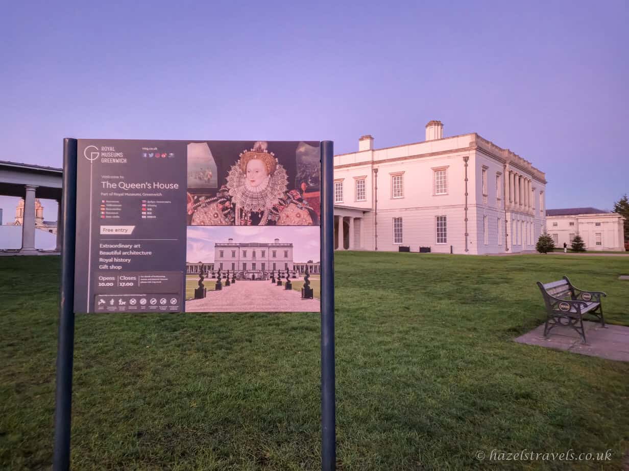 Information sign for the Queen’s House in Greenwich on a deep green lawn, with the pale cream neoclassical building behind it under a soft lilac and blue dusk sky, and a dark wooden bench to the side, London