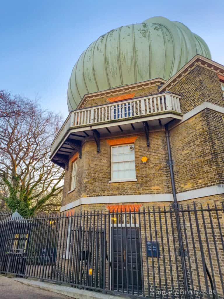 Exterior of the Royal Observatory in Greenwich with a large pale green domed roof atop a yellow-brown brick building, white balcony railings, black iron fencing, bare winter trees, and a clear light blue sky, London