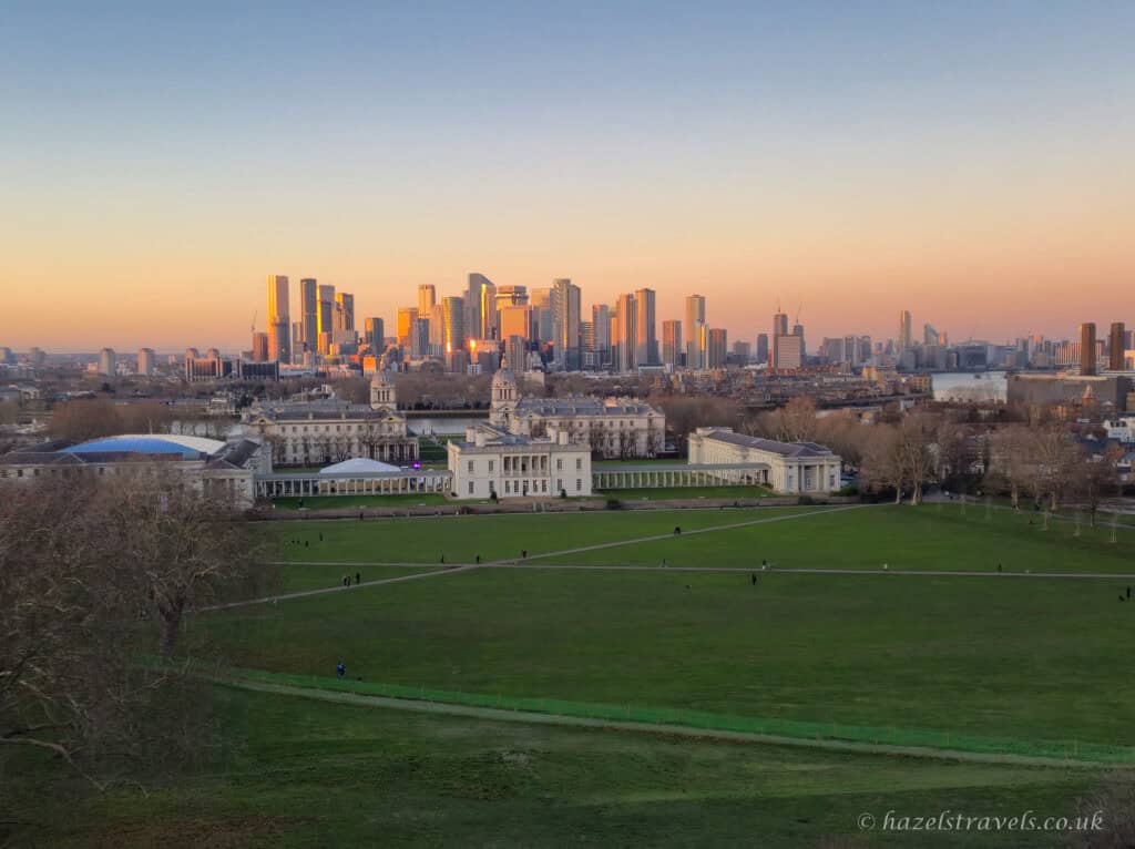 Panoramic view from Greenwich Park with deep green lawns in the foreground, the cream and white Old Royal Naval College centred below, and the Canary Wharf skyline glowing amber and gold beneath a soft peach and pale blue winter sunset sky in London