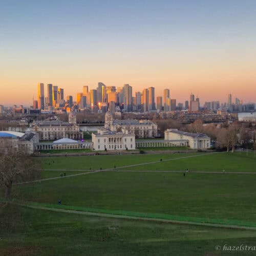 Panoramic view from Greenwich Park with deep green lawns in the foreground, the cream and white Old Royal Naval College centred below, and the Canary Wharf skyline glowing amber and gold beneath a soft peach and pale blue winter sunset sky in London