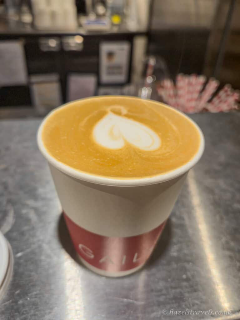 Takeaway flat white coffee in a cream and burgundy GAIL’s Bakery cup, topped with pale white heart-shaped latte art on golden-brown foam, resting on a brushed metal counter in a cosy London café