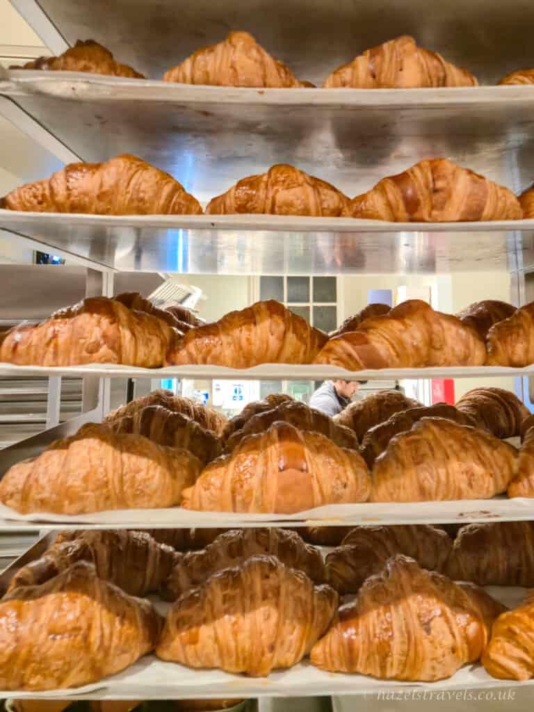 Rows of freshly baked croissants stacked on metal bakery shelves, with golden brown flaky pastry layers and a warm glow inside a London bakery