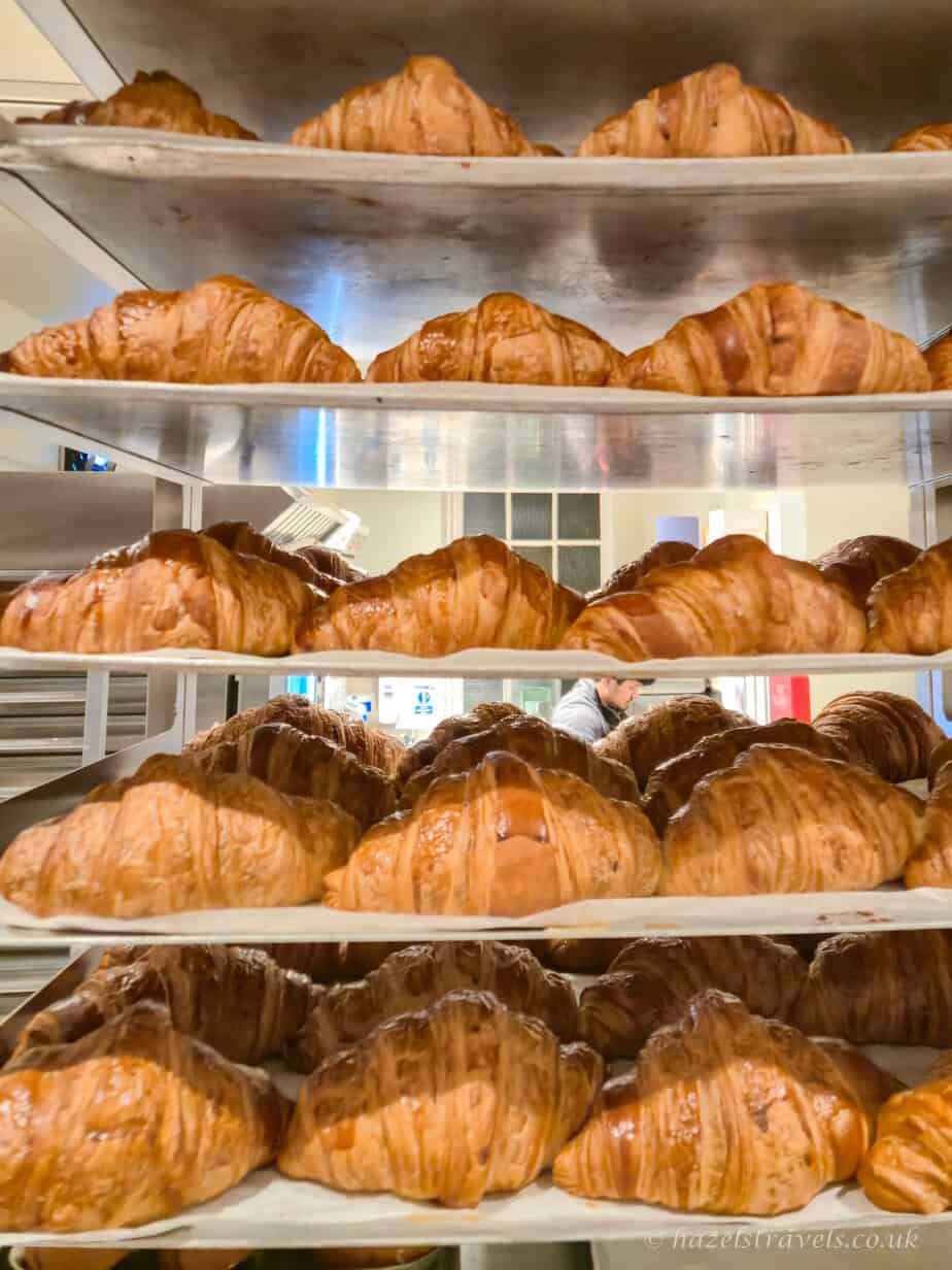 Rows of freshly baked croissants stacked on metal bakery shelves, with golden brown flaky pastry layers and a warm glow inside a London bakery