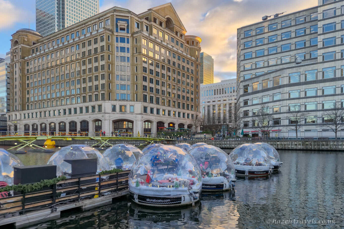 Clear plastic igloo boats floating on calm water at Canary Wharf, reflecting warm golden light from surrounding modern glass and beige office buildings under a soft pastel evening sky in London