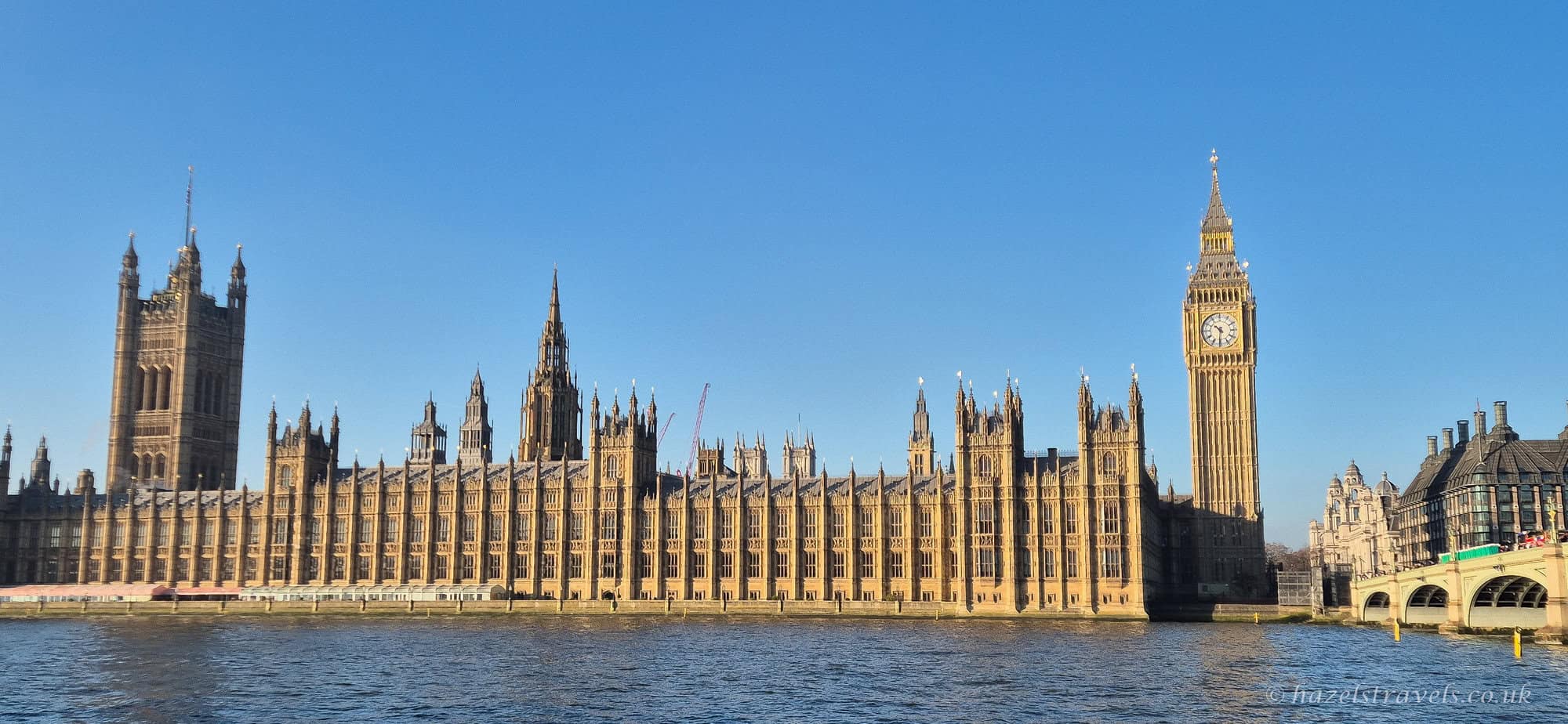 The Houses of Parliament stretching along the River Thames, with warm golden stone façades, Big Ben rising above the complex, and a clear bright blue sky reflected in the rippling water in London