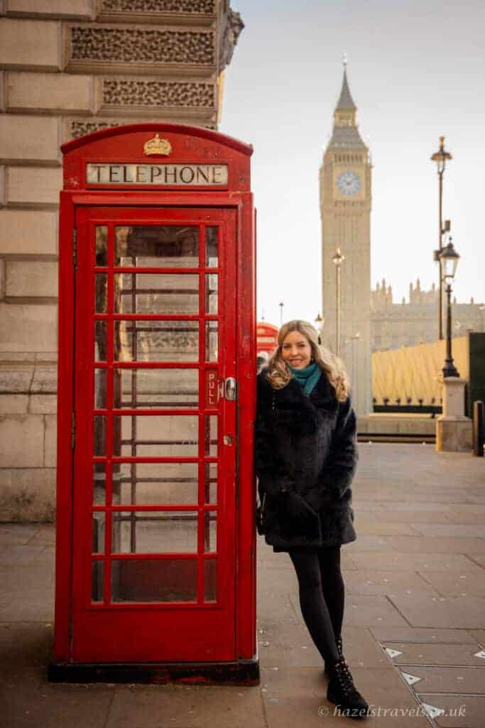 Person smiling beside a bright red London telephone box on Westminster Bridge, wearing a black coat and teal scarf, with Big Ben and the Houses of Parliament softly lit in the background under a pale winter sky