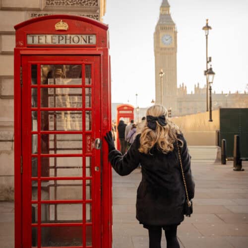 Person in a black coat standing beside a classic red London telephone box on Westminster Bridge, with Big Ben and the Houses of Parliament softly lit in the background under a pale winter sky