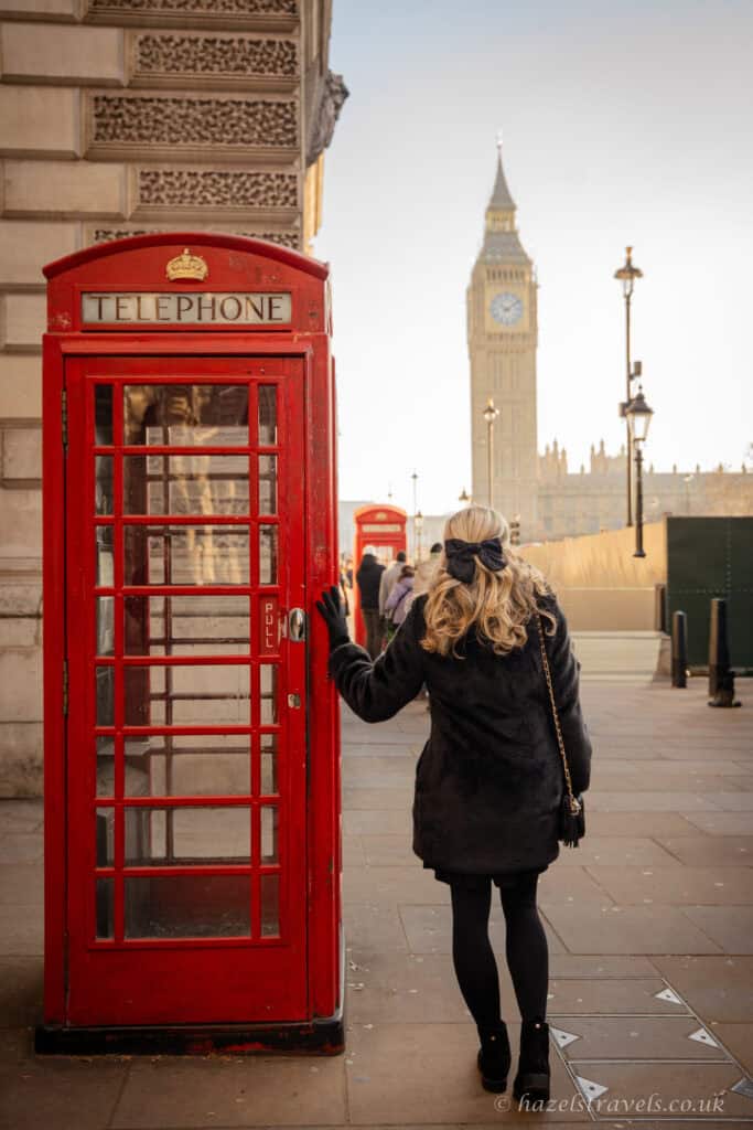 Person in a black coat standing beside a classic red London telephone box on Westminster Bridge, with Big Ben and the Houses of Parliament softly lit in the background under a pale winter sky