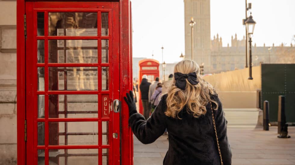 Person in a black coat standing beside a classic red London telephone box on Westminster Bridge, with Big Ben and the Houses of Parliament softly lit in the background under a pale winter sky