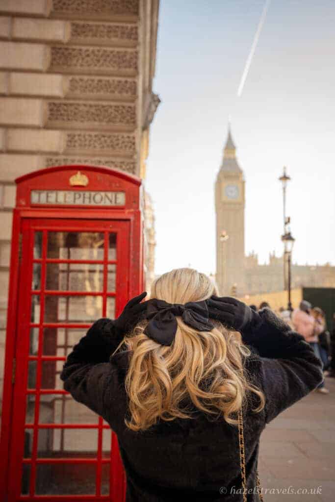 Person with blonde hair tied in a black bow standing beside a bright red London telephone box, facing Big Ben and the Houses of Parliament in soft golden winter light under a pale blue sky