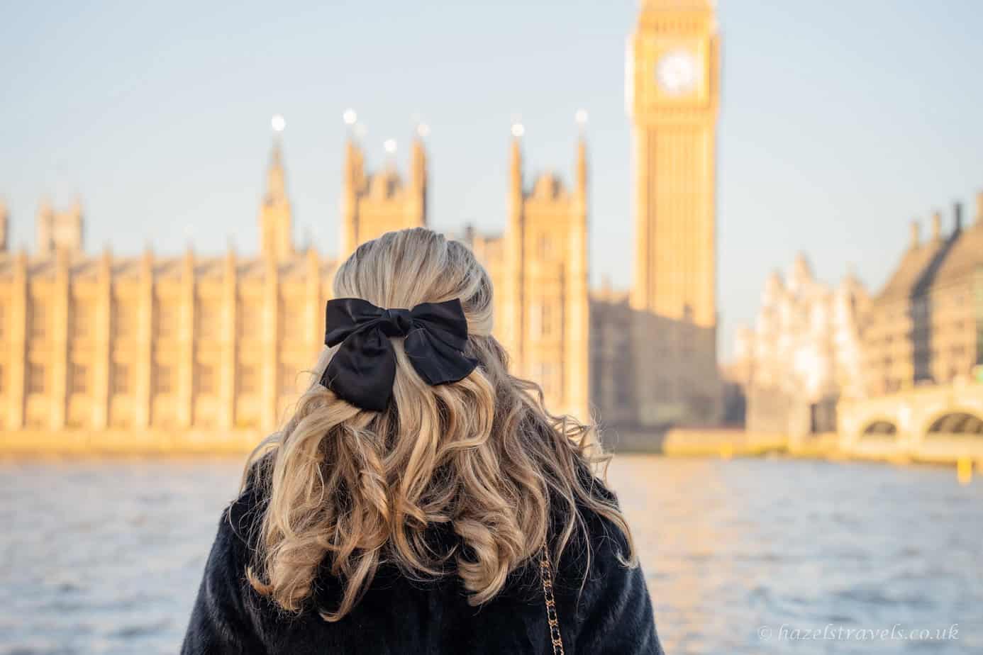 Person with blonde hair tied in a black bow standing by the River Thames, facing Big Ben and the Houses of Parliament glowing warm gold in low winter sunlight, with pale blue sky and rippling grey-blue water in London