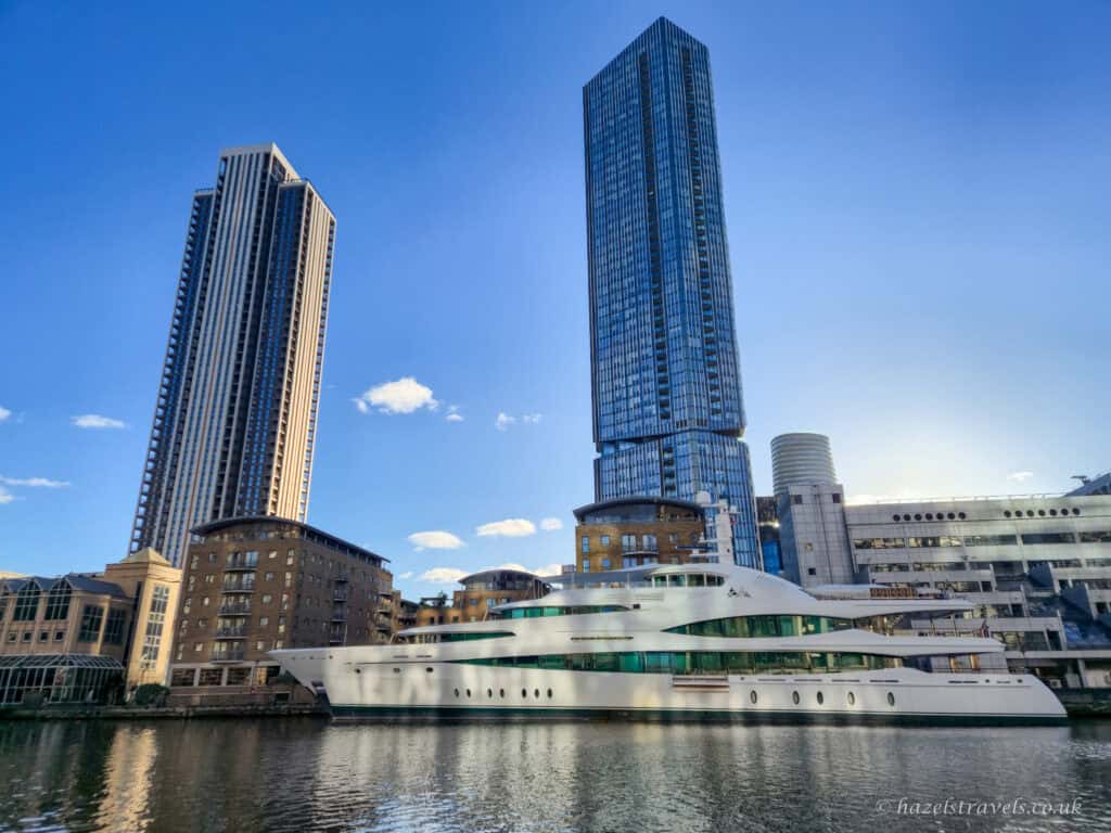 Modern waterfront scene in Canary Wharf with two tall glass skyscrapers rising into a clear blue sky, reflected in calm dock water. A large white luxury yacht is moored in the foreground, with mixed modern and brick buildings lining the water’s edge.